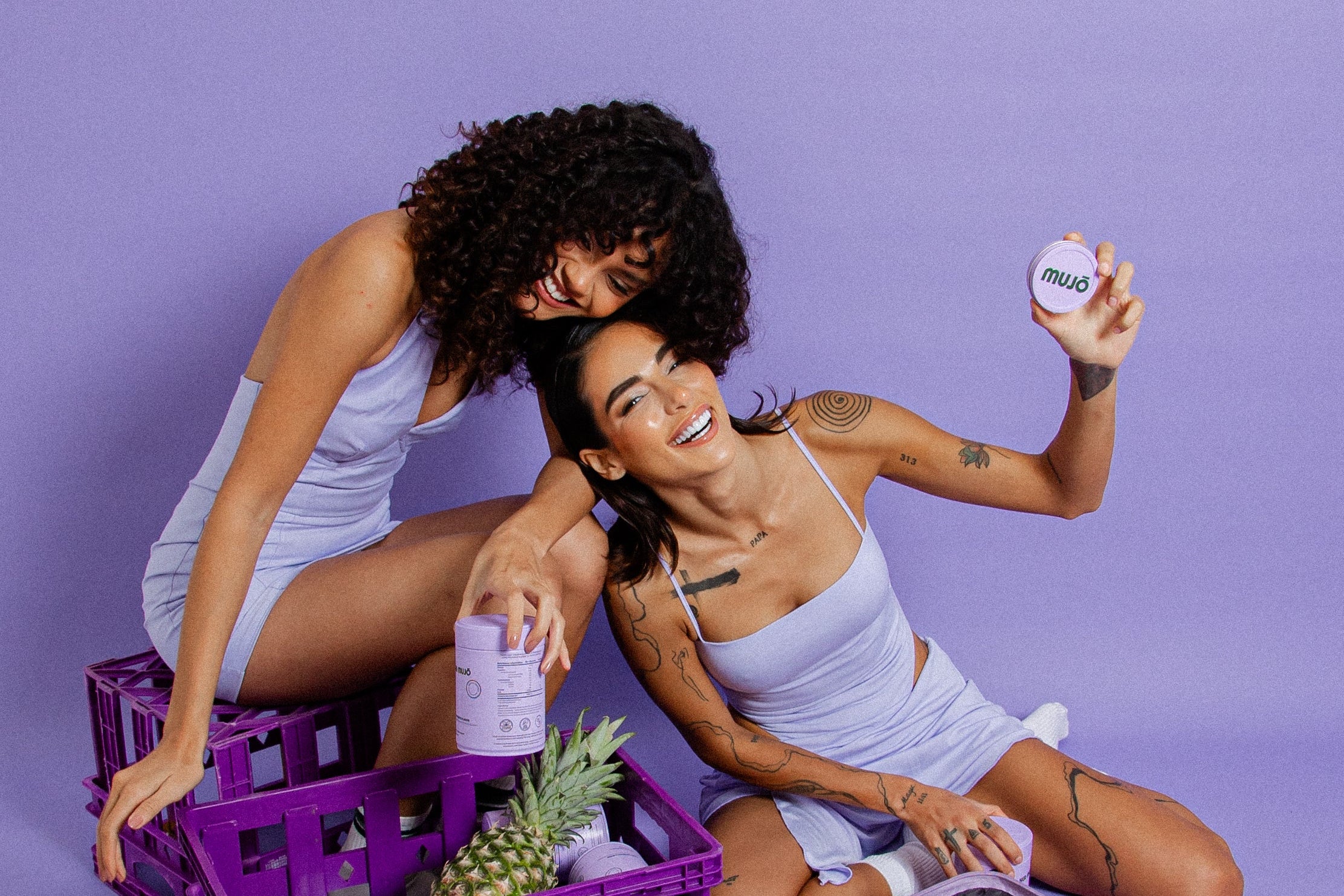 Two women with a suitcase full of MUJO jars on a purple background